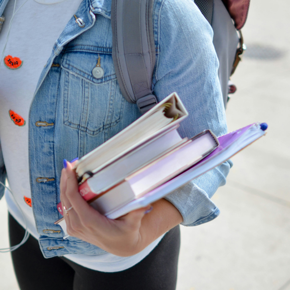 Persona che trasporta una pila di libri mentre indossa una giacca di jeans e uno zaino, apparentemente pronta a "richiedi un contributo" per il prossimo grande progetto.