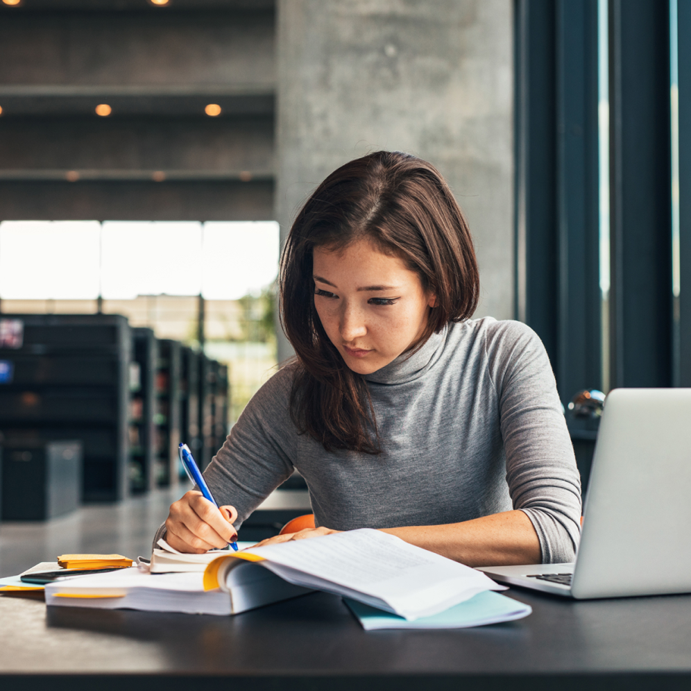 una donna studia diligentemente alla sua scrivania con un libro aperto e un computer portatile. La sua attenzione è molto elevata.